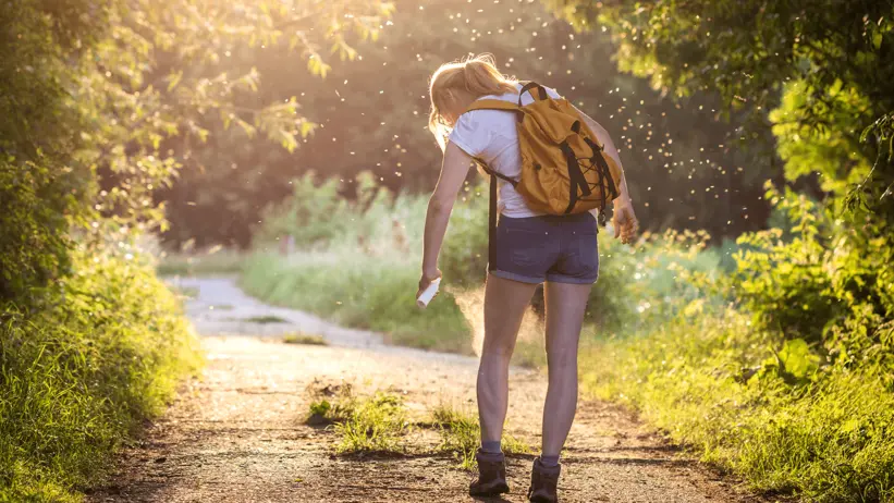 A woman with a yellow backpack stands on a sunlit path, using insect spray, to try and prevent insect bites. She's surrounded by lush greenery, conveying a peaceful outdoor setting.