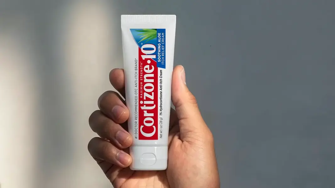 A hand holds a tube of Cortizone-10 cream against a plain gray background. The tube's red and blue label stands out, suggesting soothing relief.