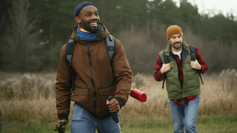 The image features two men walking through a grassy field, each carrying a backpack and a ski pole.