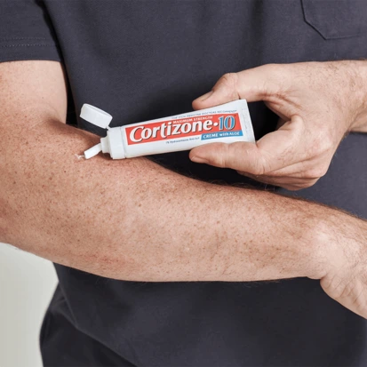 A person applies Cortizone-10 cream onto their forearm. The tube is held in one hand, cap open, ready to soothe and relieve skin irritation.
